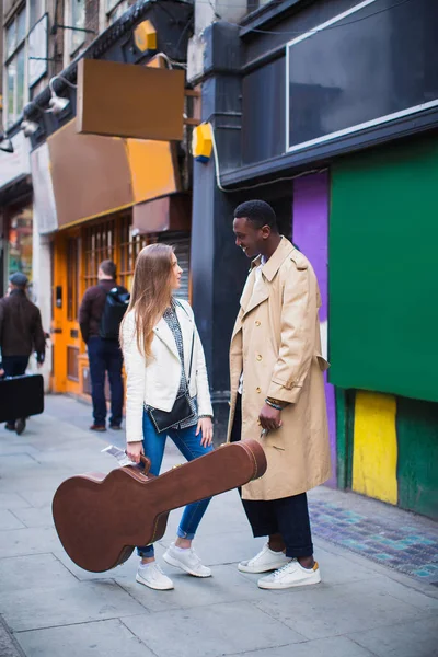 Two young people stand and talk, one of them keeps guitar case in hand, outdoor