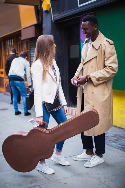 Two young joyful people stand and talk, one of them keeps guitar case in hand, outdoor