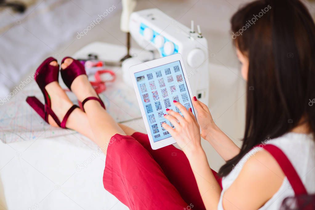 Female brunette sits and keeps her legs on a table, keeps the tablet in her hands and looks at in, in atelier