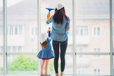 Back view of young mother who washes the windows and her little daughter helps mother indoor clipart