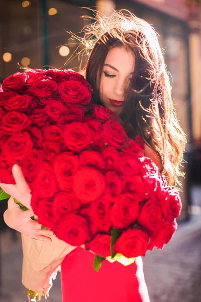 Elegant lady stands and keeps big bouquet of red roses in hands, on the street
