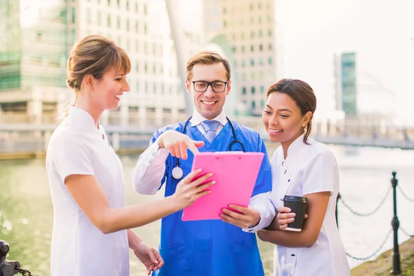 Happy smiling doctor and two nurses look at folder and discuss something during coffee break, outdoors