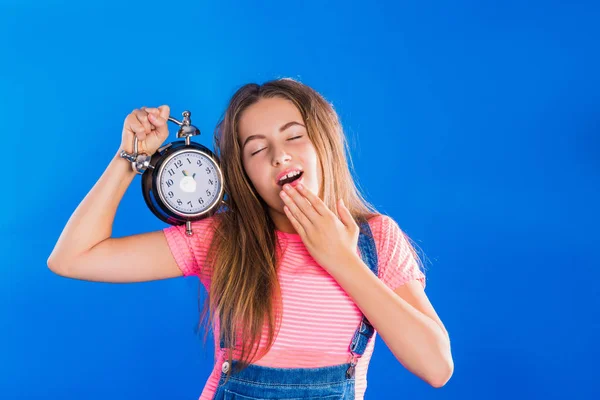 Portrait of young sleepy woman who stands on blue background and keeps alarm clock