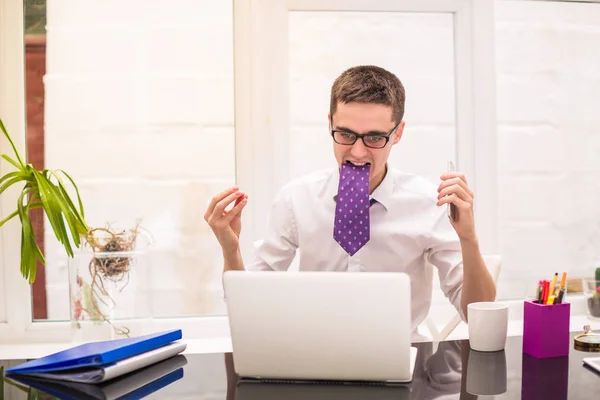 Young businessman looks at laptop, keeps necktie in teeth and keeps the phone in hand at the office