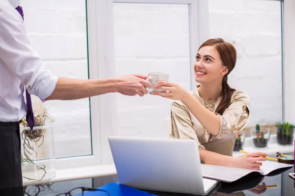 Office manager gives cup of coffee to young cheerful businesswoman who sits at the table and does her affairs at the office