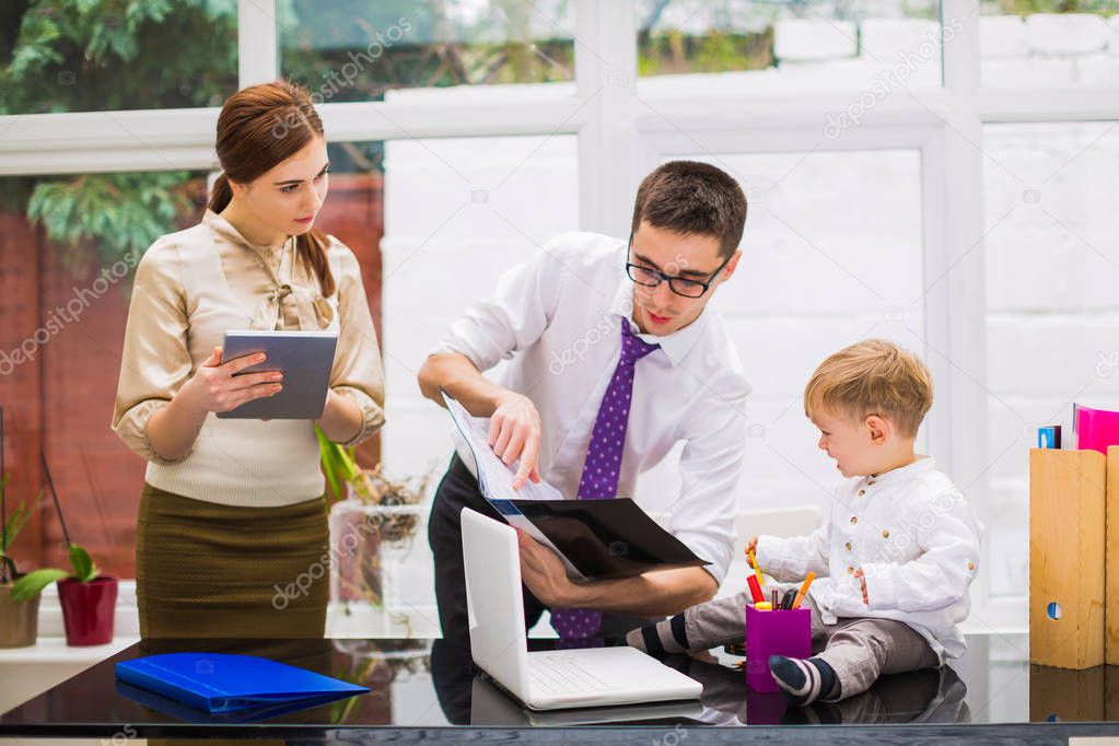 Young father shows an opend folder to his little son who sits on office table, assistant stands near them with the tablet in hands, at the office