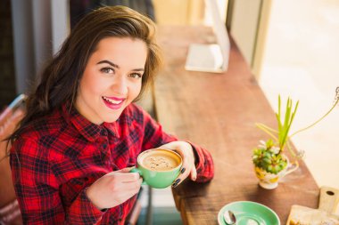 Neşeli kız öğrenci pencerenin yakınındaki masada fincan kahve ile onun elinde zaman ara, kafede oturuyor