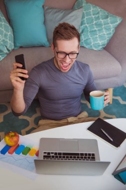 Genç başarılı adam yere evde oturma ve çalışma ve telefonu ve fincan elinde tutan ve joying