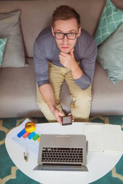 Evde kanepede oturan ve çalışan ve telefon elinde tutan genç düşünen adam