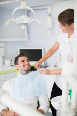 Görüntü bir kadın dişçi klinikte tedavi öncesi erkek hasta önlük ayarlama.