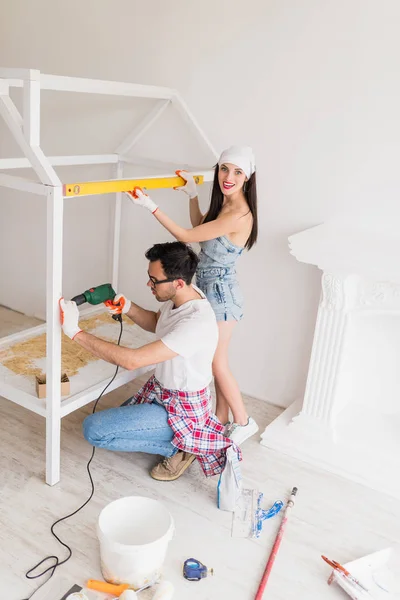 The young couple repairs crib at home, smiling woman keeps meter and measures crib, a serious man drills deer for dowels in crib