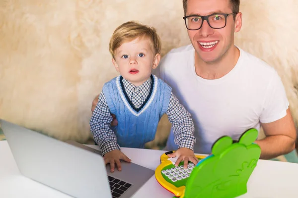 年轻快乐的父亲和他的小儿子坐在桌旁 在室内使用笔记本电脑 — 图库照片