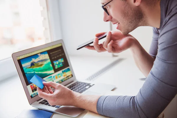 The young man working at home, he speaking by phone, looking the laptop and holding business card in hand