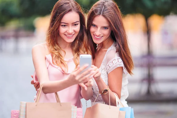 两个年轻微笑的女人的肖像 她们拿着购物袋站在大街上 在电话里做自拍照片 — 图库照片
