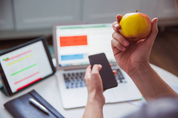 Крупный план яблоко в руке и телефон в другой руке на фоне работы вещи на столе в помещении

