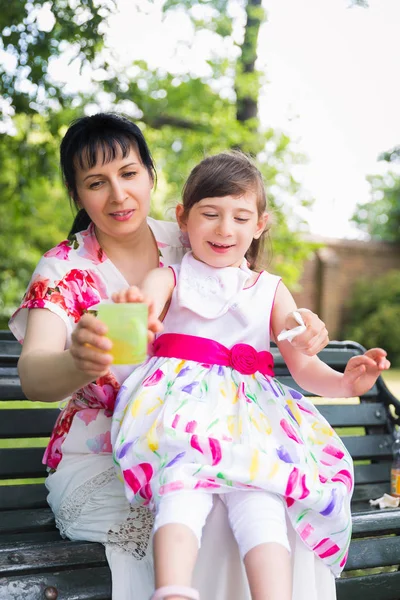 屋外のベンチに座っていると保つラップで無効とドリンクの母維持カップを手に 彼女の美しい娘 若い母親 — ストック写真