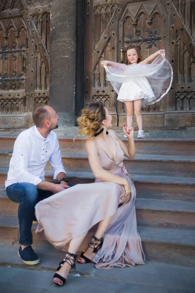 The young happy parents sitting on the stairs of old building outdoor and looking at their little cute daughter who standing on the stairs and holding by dress