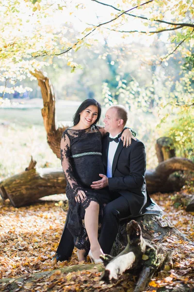 A pregnant cheerful woman sits on lap of her husband who keeps his hand on her belly in the park