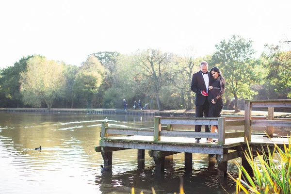 若い妊婦は抱擁で彼女の幸せな夫と池のそばに立つし 彼は手に小さな赤い靴を保持 — ストック写真