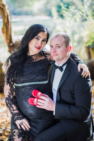 A pregnant divine woman sits on lap of her husband who keeps little red shoes on belly of his wife in the park