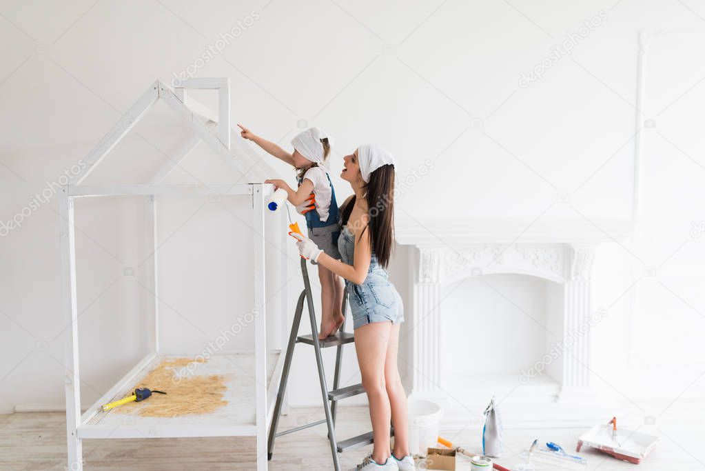 The young smiling mother does repair at home, she stands on ladder near her daughter and they draw crib with help of a platen and girl shows forefinger on roof of crib