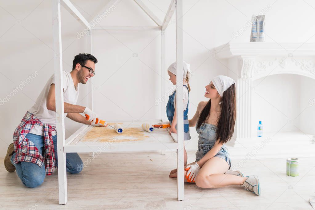 The young happy family does repair at home, smiling father and his daughter paint the crib with help of the platens, cheerful mother sits near daughter
