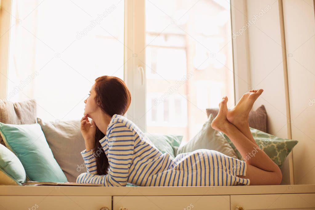 Side view of young beautiful woman who lying on the sofa in the room and looking in the window