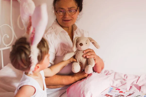 Happy grandmother sitting on a bed with her little grandson who is in the bunny ears and she holding a toy in a room at home