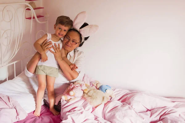 うさぎの耳は 彼女の最愛の小さな孫に近いおもちゃでベッドに座っていると寝室で彼を抱きしめる幸せの祖母 — ストック写真