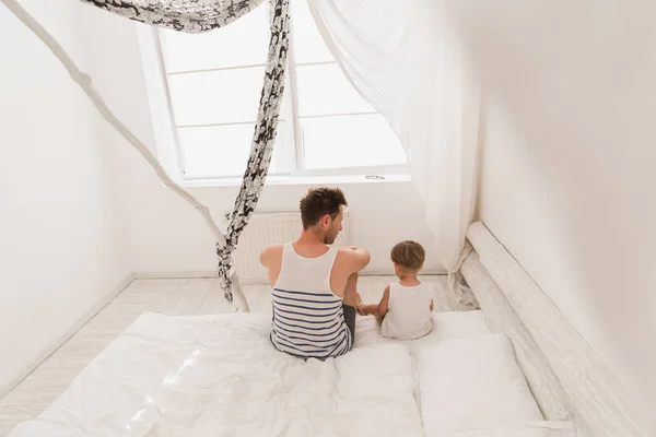 stock image Back view of the young father with his little son  are sitting on a bed in a white room