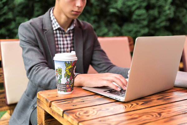 Крупный план молодого человека, сидящего за столом на открытом воздухе и работающего с ноутбуком и пьющего кофе
