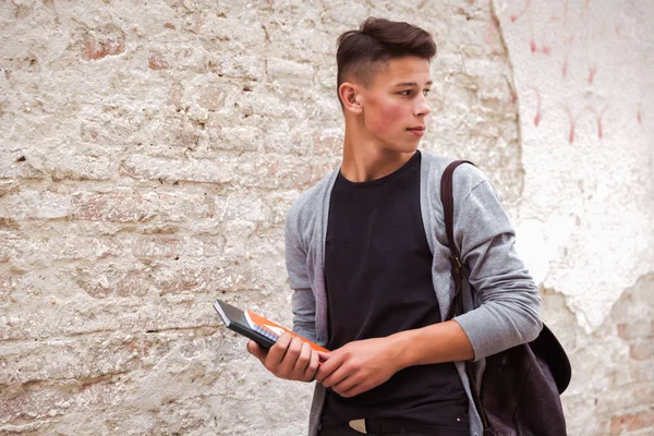 Portrait of the young man who is standing outdoor near the old house wall and holding the notebooks in hands and looking back