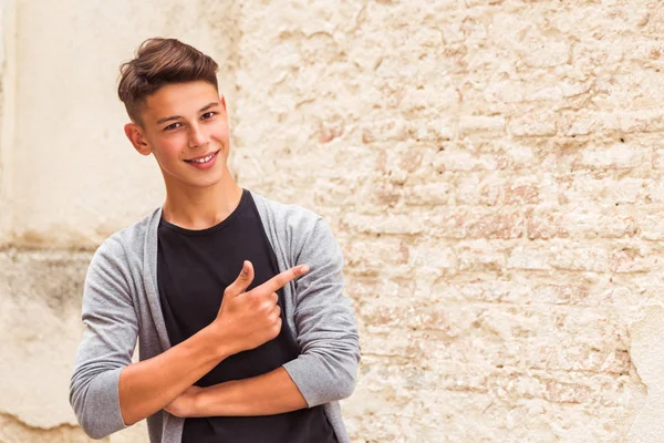 屋外古い家の壁の近くに立って 人差し指で脇を示す幸せな若い男の肖像 — ストック写真