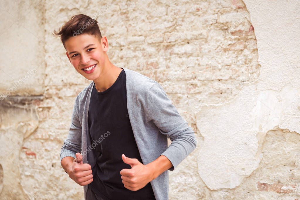Portrait of the happy young man who is standing outdoor near the old house wall and showing thumbs up