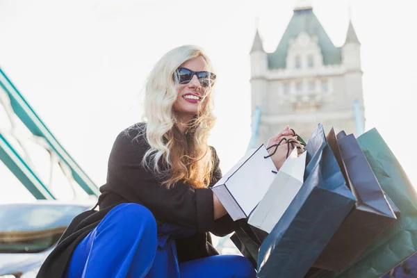 微笑的美丽的年轻金发碧眼的女人坐在桥上 手里拿着购物袋 — 图库照片