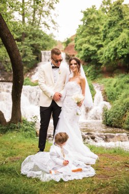 The happy newlyweds standing together against the background of a waterfall and cute little boy sitting on a dress of the bride near them clipart