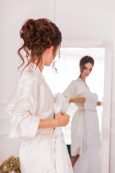 美しい若い花嫁を屋内の鏡の前に立っていると彼女のブライダル ガウンを保持 — ストック写真