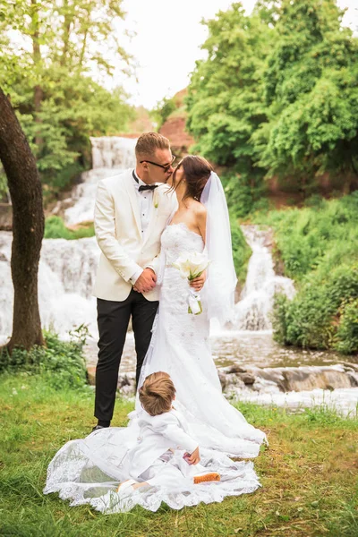 幸福的新婚夫妇在瀑布的背景下接吻 一个小男孩坐在新娘的衣服上 坐在他们旁边 — 图库照片