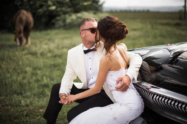 美しい新婚屋外馬の近くの古い車のバンパーに抱擁で座っているとキス — ストック写真