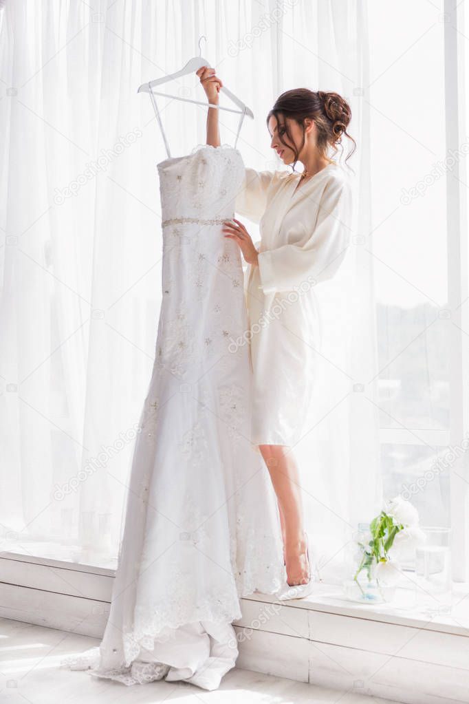 The happy, beautiful bride standing near the window indoor and holding her bridal gown and looking at it