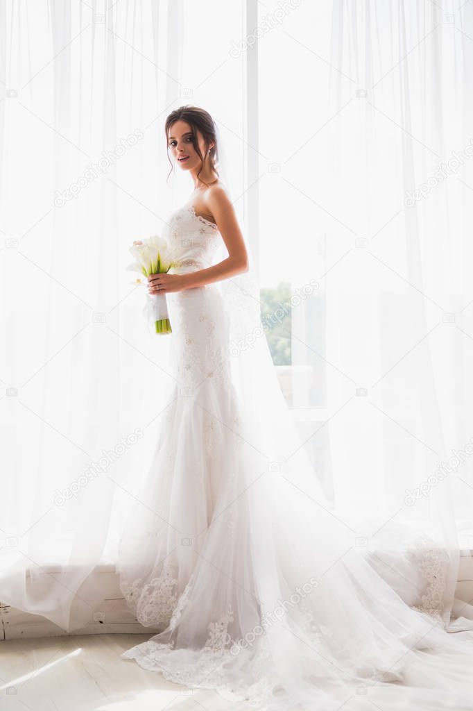 The beautiful happy bride standing near the window indoor and holding a bouquet in hands