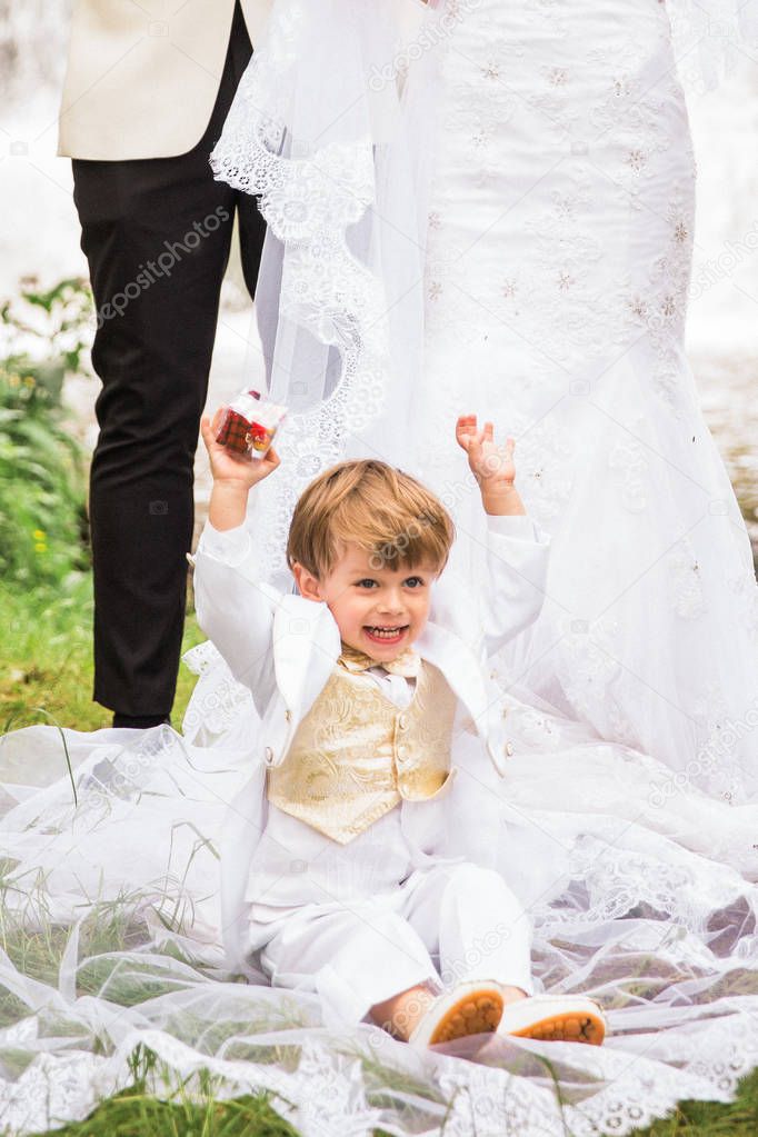 The joyful little boy sitting on a train of the bridal gown of a bride outdoor