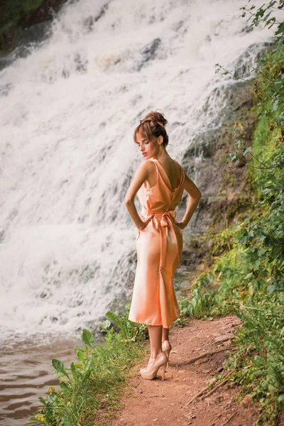 The beautiful young lady in dress standing near the waterfall