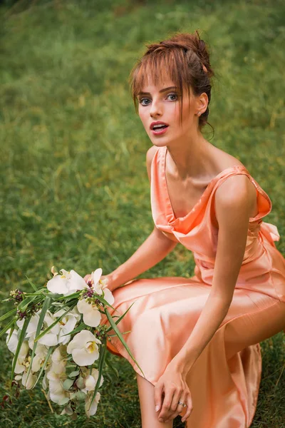 The beautiful young lady in the dress sitting on the grass and holding flowers in hand