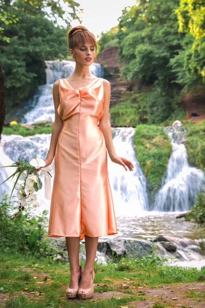 The beautiful young lady in the dress standing against the background of the waterfall and holding the flowers in hand and looking down