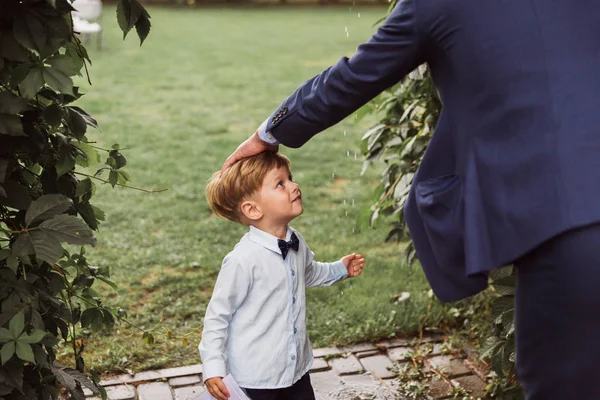 Side view of the little cute boy who is standing outdoor and looking at his father who is stroking him on the head