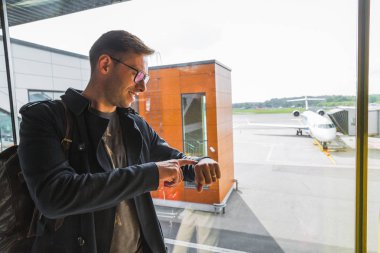 Havaalanında pencerenin yanında duran ve eline bakan gülümseyen genç adamın yan görünümü