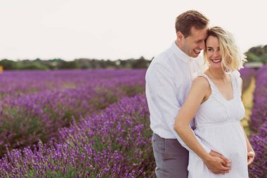 Mutlu genç adam lavanta tarlasında neşeli hamile karısına sarılıyor.