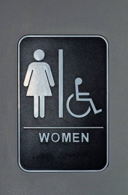 Banyo duvarındaki dikey tabela, kadınları ve engelli kadınları sadece o banyoya atamaktadır. Tabelanın altındaki braille kullanımını gösterir.