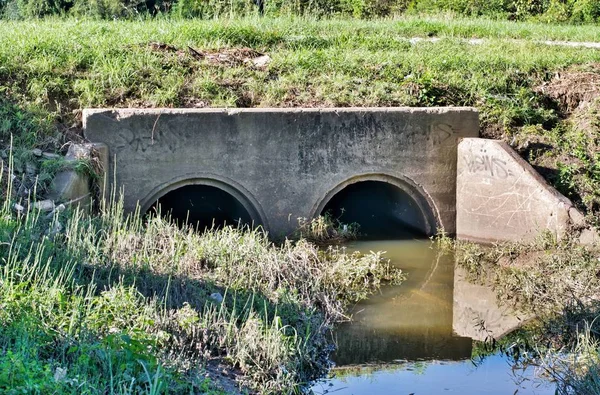 Culvert with twin concrete drains under an unpaved road in Humble, Texas. Dirty polluted water is stagnant in the drainage ditch with some trash lying in the grass. City pollution is getting worse.